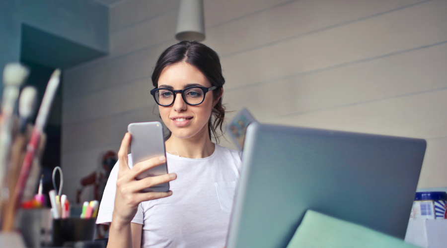 women looking at her phone and sitting in front of a laptop with pens and art supplies on her desk