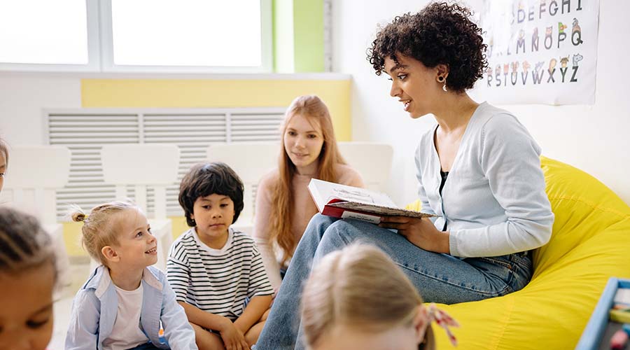 An early years educator reads to children while sitting on a bean bag. A student sits with the children.