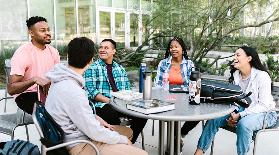 5 T Level students sit around a table, in the courtyard of a college. They are smiling