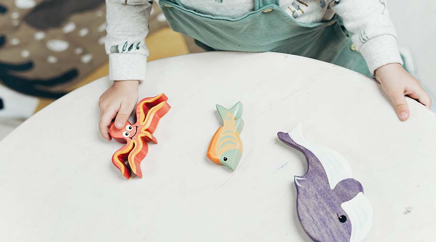 Three wooden toys are on a table, a fish, an ocotupus and a whale. A young child's hand holds the octopus toy.