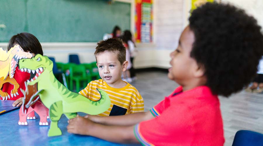 Two young boys play with cardboard dinosaurs in a classroom.