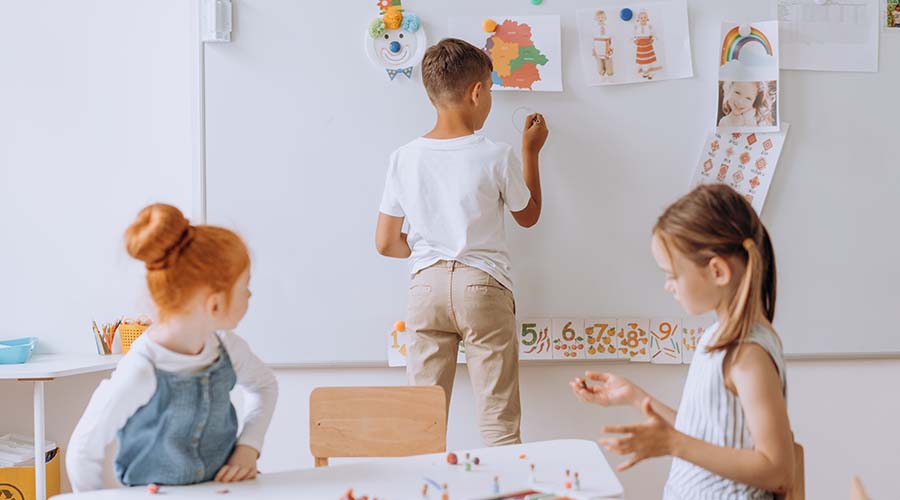 three children play in a pre-school. A boy os drawing on a whiteboard while two girls sit at a table.