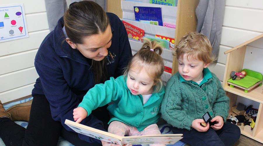 An Early Years teacher and two children in green uniform share a book.