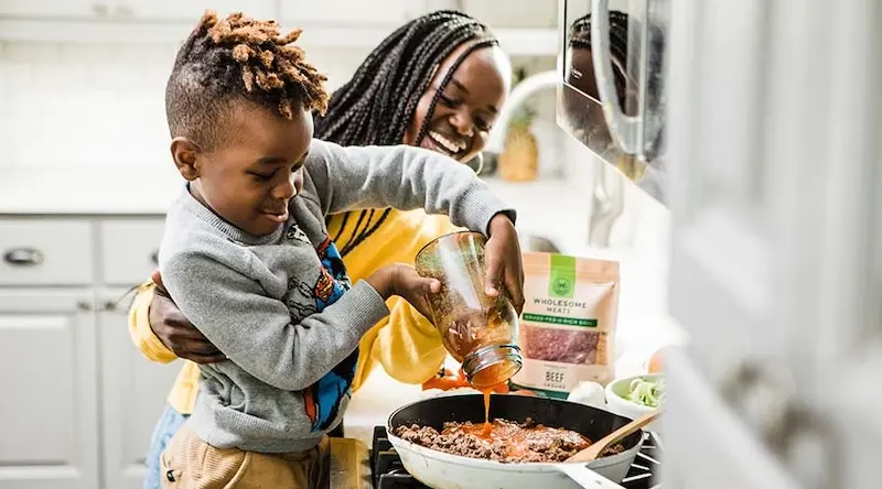 A woman supports a child under his arms, as he pours sauce into a pan of food, in a kitchen