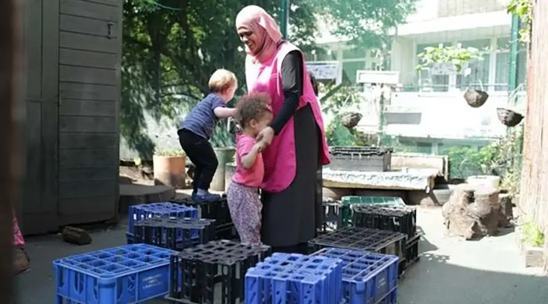 An educator holds hands with a little girl as she balances on milk crates. A little boy is also climbing on the milk crates behinds them.