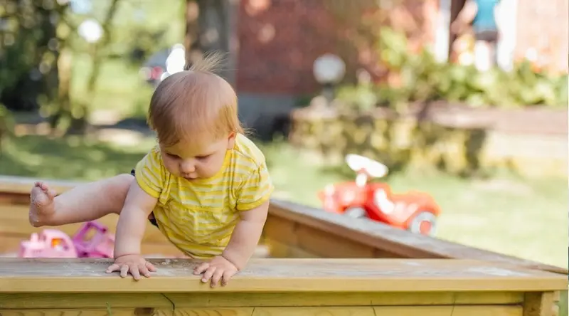 Toddler in a yellow striped shirt climbing on a wooden playground structure with toys in the background