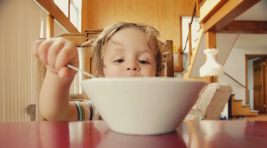 A young child with light hair eagerly reaches for a spoon in a white bowl, seated at a red table. The warmly lit room feels cozy and inviting.