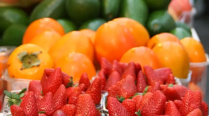 Fresh strawberries displayed in the foreground, with vibrant orange persimmons and green avocados in the background, conveying a sense of abundance.