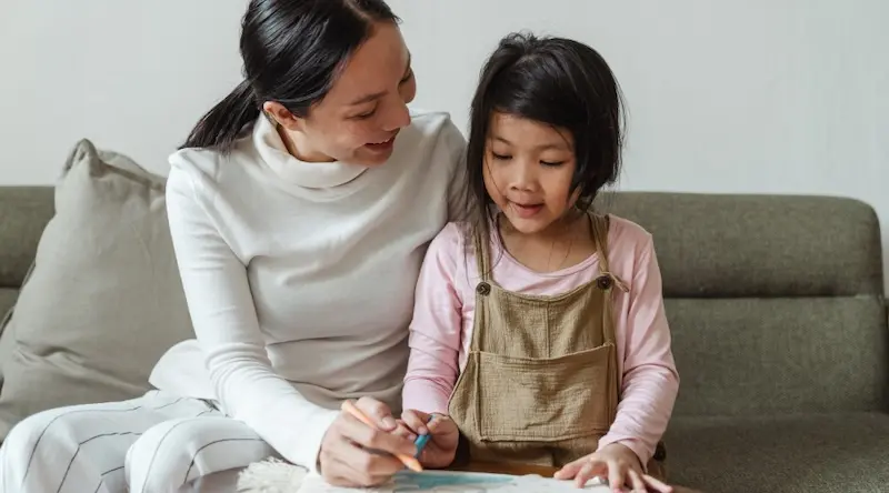 A woman and child sit on a sofa, smiling and drawing together. The woman is wearing a white turtleneck, and the child wears a pink shirt with overalls. The scene feels warm and nurturing.
