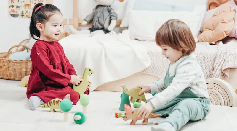 Two children sit on a light-colored floor, joyfully playing with toy dinosaurs. The girl in a red dress smiles, while the boy in green overalls is focused. A cozy room with soft furnishings enhances the playful, warm atmosphere.
