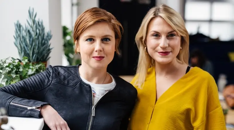 Two women smiling indoors, one in a black jacket and the other in a yellow top. They stand near plants, creating a warm, friendly atmosphere.