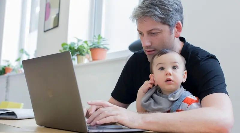 A man in a black shirt works on a laptop at a home desk, with a curious baby on his lap. The scene is bright, with potted plants on the windowsill.