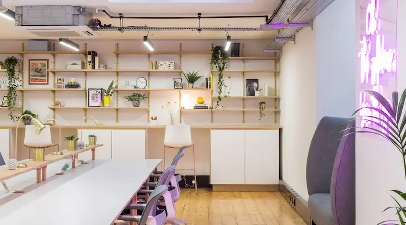 Bright, modern office space with long white tables, pastel chairs, and wall shelves adorned with plants, books, and decor. Neon light adds a warm glow.