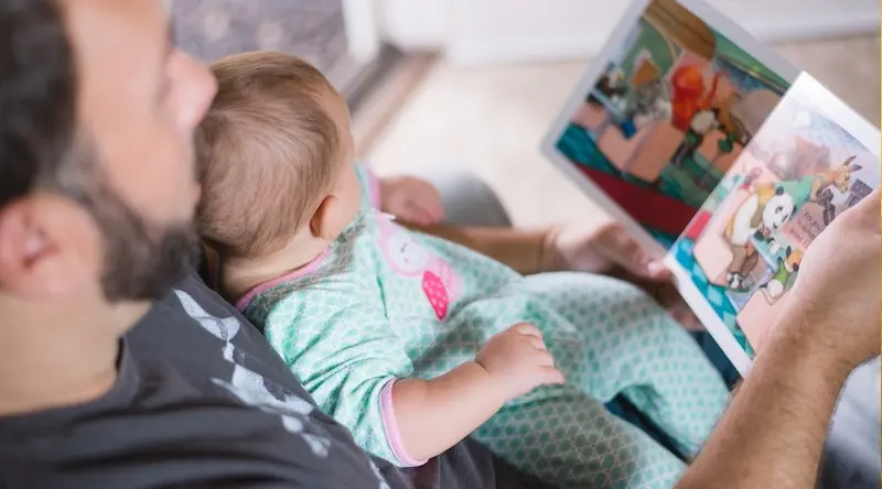 A man reads a colorful children's book to a baby on his lap. The baby wears a pastel onesie with a cute print. The scene feels warm and nurturing.