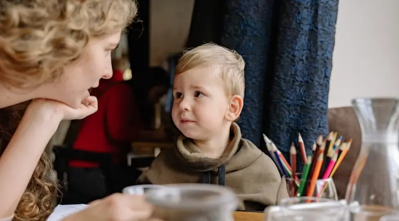 A woman with curly hair and a young boy share a tender moment at a table. Colored pencils and a carafe are nearby, creating a warm, creative atmosphere.