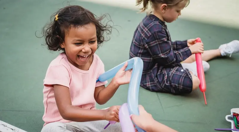 A joyful child in a pink shirt laughs while holding colorful balloon animals outdoors. Another child in plaid sits nearby, focused on a red balloon.