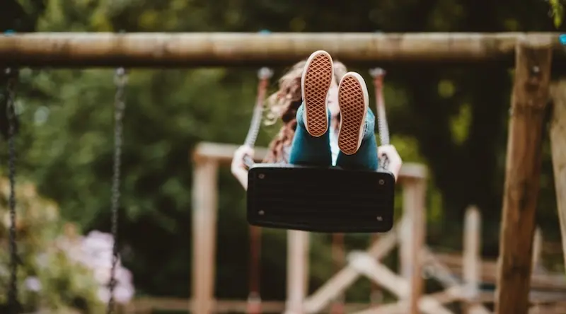 Child in a red jacket reaches up to grab a large stick suspended in a forest, surrounded by tall trees. The scene conveys adventure and playfulness.