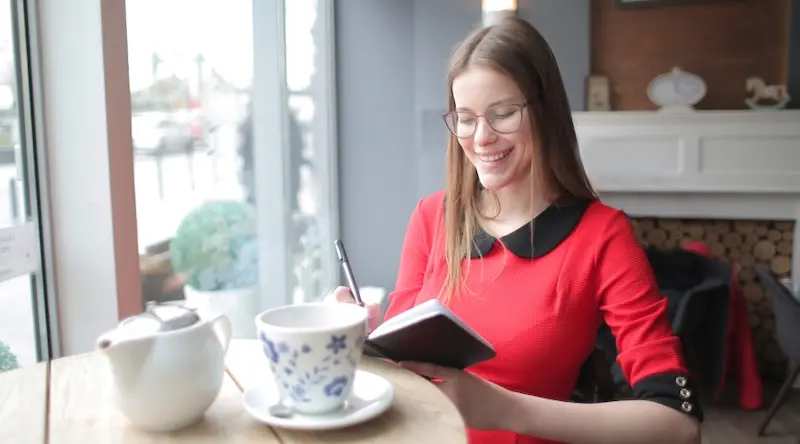 A smiling woman in glasses and a red dress writes in a notebook at a café. A teapot and floral cup are on the table, creating a cozy atmosphere.