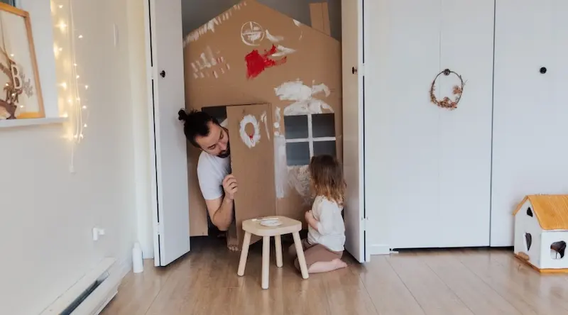 A man peeks from a cardboard playhouse painted with red and white, while a child sits painting. They appear engaged and playful in a cozy room.