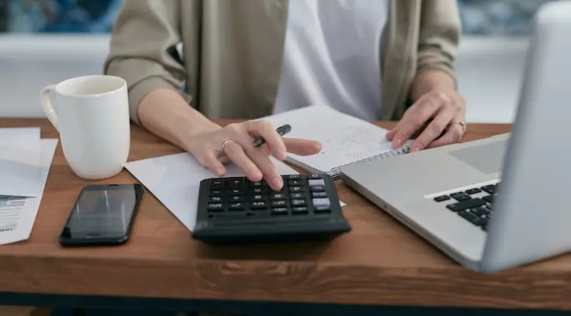 A person uses a calculator on a wooden desk, surrounded by a laptop, smartphone, coffee mug, and papers, conveying a focused and productive work environment.