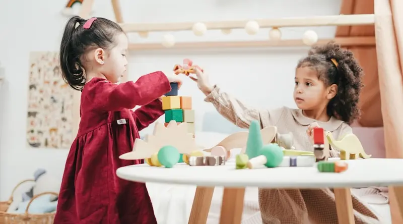 Two young girls play with colorful wooden blocks at a small table. One wears a red dress, the other beige. The scene is lighthearted and engaging.