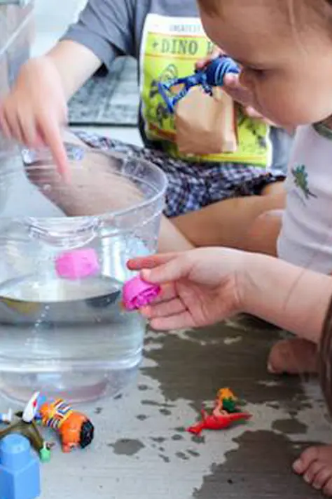 A child with a clear bowl of water. Another child is putting a pink bottle cap in the water.