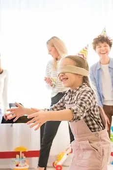 A child wearing a blindfold and plaid shirt plays pin the tail at a party. Other children in hats laugh and cheer in a bright room filled with toys.