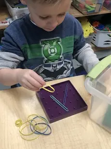 A child in a superhero shirt plays with rubber bands on a purple pegboard at a table, focused and engaged. Bins of toys are in the background.