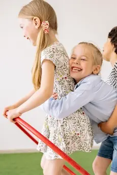 Three children playfully ride a red hula hoop in a bright room. The front child is smiling, exuding joy and energy, creating a happy, playful atmosphere.