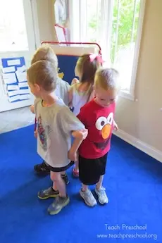 Children stand in a circle on a blue carpet, back-to-back, in a classroom with bright windows. One child wears a red Elmo shirt, creating a playful atmosphere.