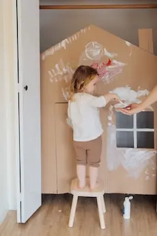 A child stands on a stool painting a cardboard house with white paint, creating an artistic and playful scene. An adult hand offers more paint.