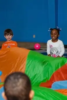 Children smile and laugh as they hold a colorful parachute indoors. A pink ball is nearby, creating a joyful, playful atmosphere against a blue wall.