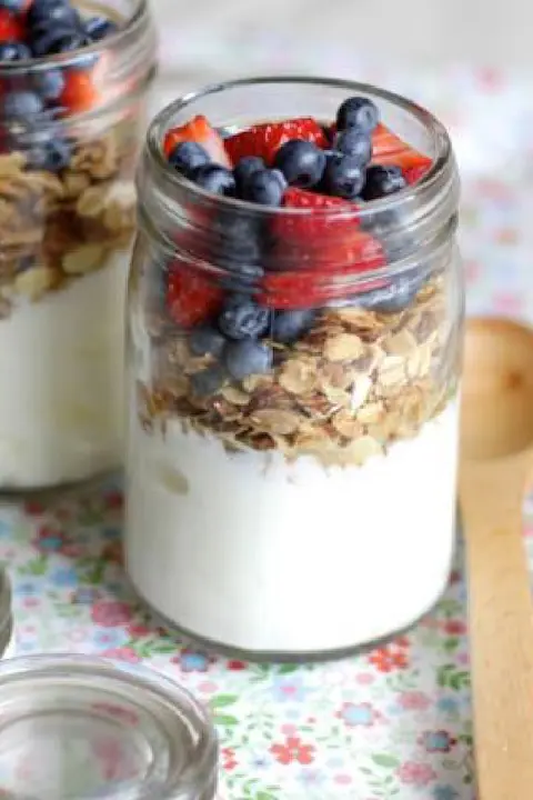 Jar filled with creamy yogurt, a layer of granola, and topped with fresh blueberries and strawberries. It sits on a floral tablecloth beside a wooden spoon.