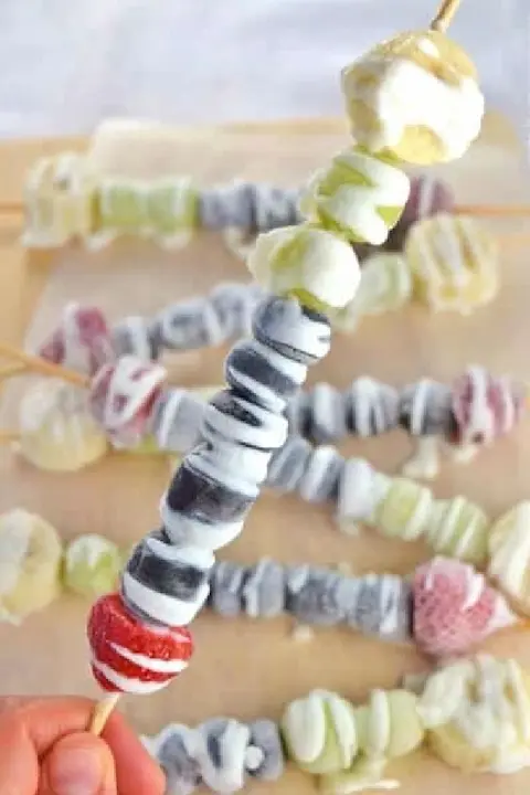 Skewers of assorted frozen yogurt-coated berries and fruits, including strawberries, grapes, and bananas, on a wooden board. Fresh and colorful.