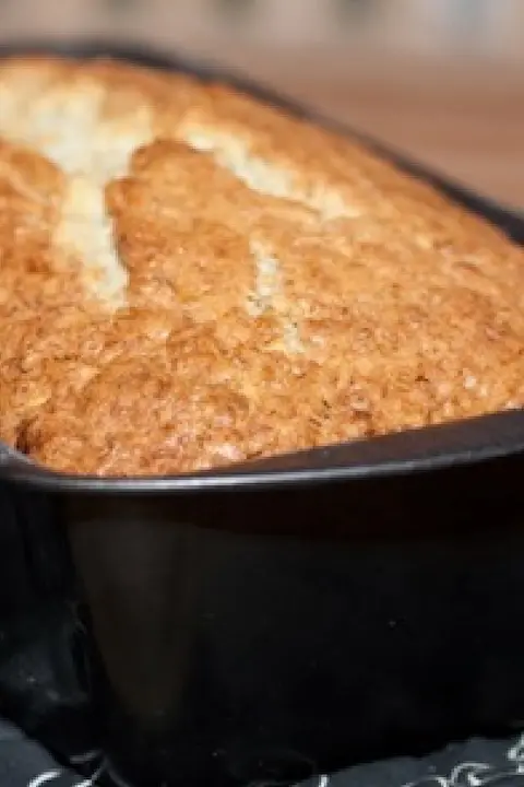 Golden brown bread loaf baked in a black rectangular pan, with a crisp, textured crust. The warm tones suggest a cozy and inviting homemade feel.