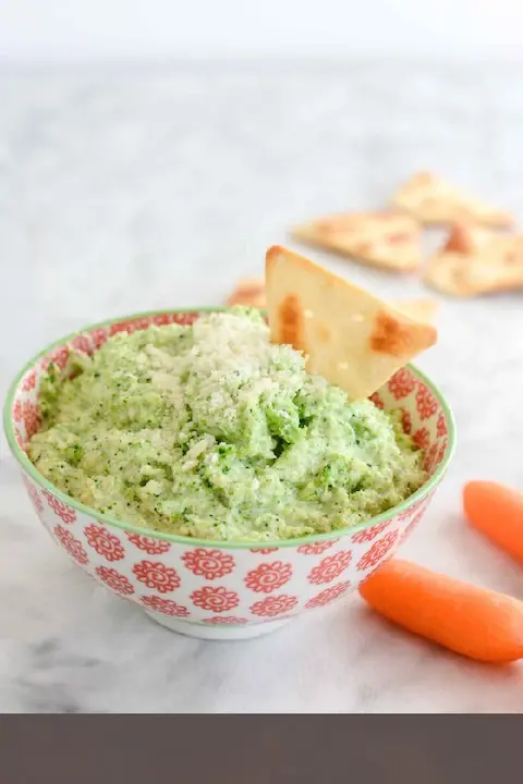 A bowl with colorful floral patterns holds a creamy green dip topped with shaved cheese and a tortilla chip. Baby carrots and tortilla chips are nearby.