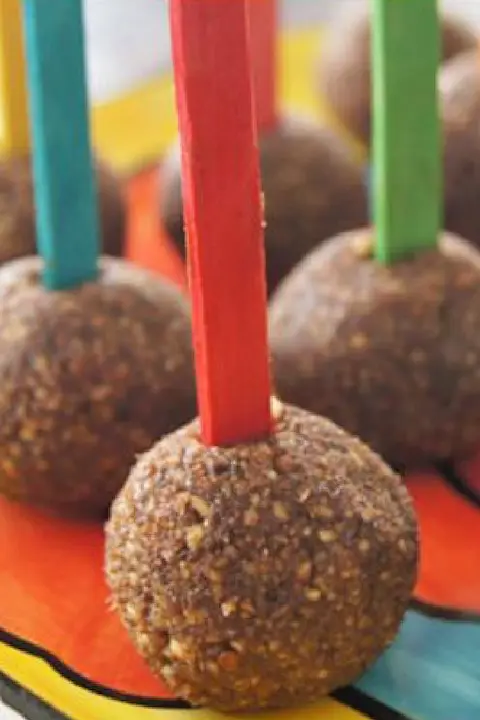 Close-up of chocolate-coated cake pops with multicolored sticks arranged on a vibrant, circular tray. The scene conveys a playful, festive mood.