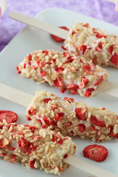 Three yogurt popsicles coated with granola and strawberry pieces on a white tray, set against a light purple background, create a fresh, summery feel.