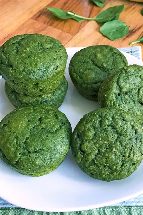 Alt text: A plate of vibrant green muffins on a wooden table. The muffins are fresh and stacked neatly, with spinach leaves scattered in the background.