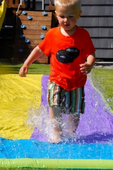 A young child with blonde hair joyfully splashes on a colorful slip 'n slide, wearing a red shirt and striped shorts. The scene is bright and playful.