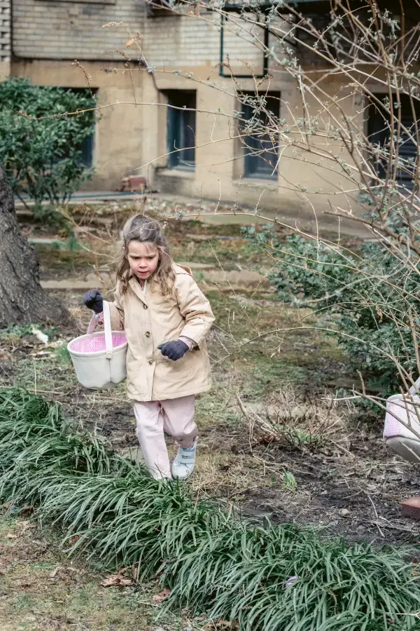A young girl in a beige coat and gloves holds a basket, exploring a garden with bare branches. She appears curious against a backdrop of greenery and a brick building.