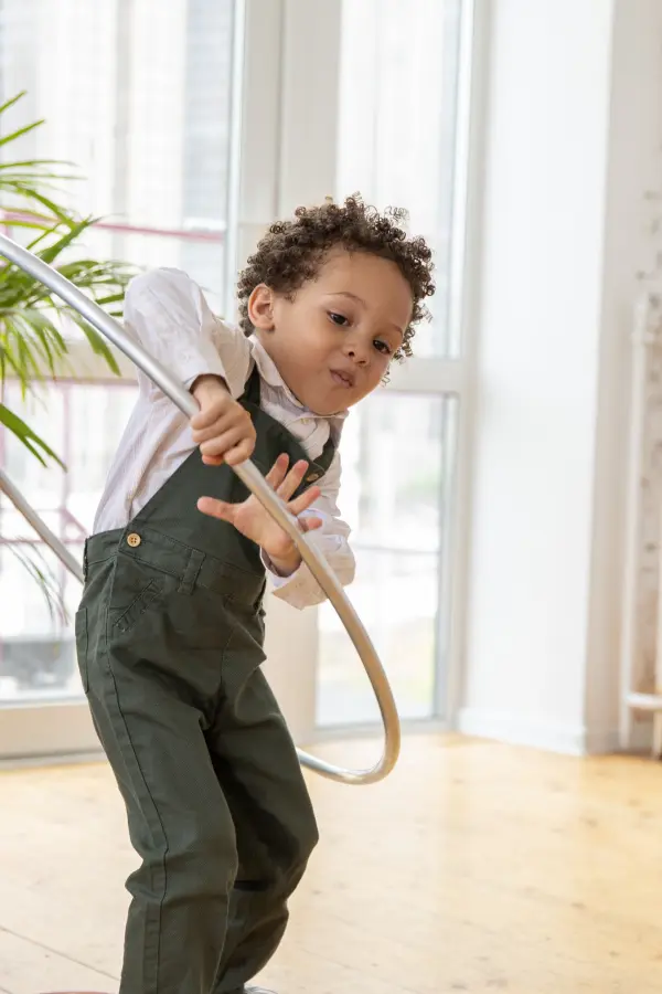 A young child with curly hair, wearing green overalls and a white shirt, joyfully plays with a hula hoop indoors. Bright natural light and a plant are in the background.