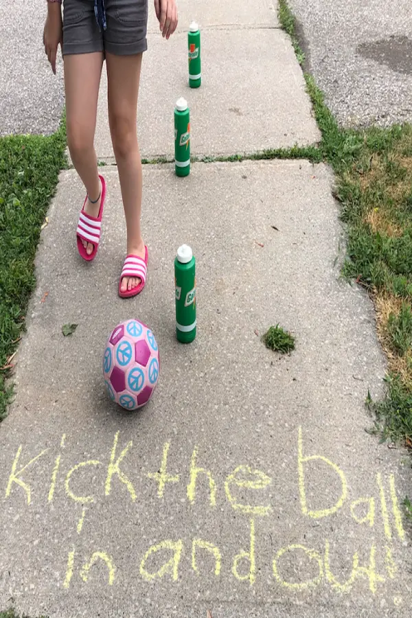 A child in pink sandals and gray shorts kicks a colorful ball between green bottles on a sidewalk. Chalk writing reads, "Kick the ball in and out."