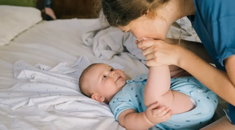 A woman gently kisses a smiling baby's foot as they lie on a bed. The scene conveys warmth and affection, with soft bedding in the background.