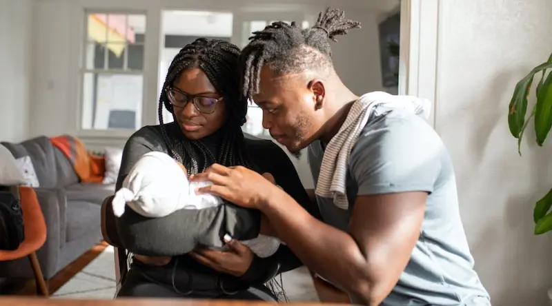 A couple lovingly gazes at their newborn wrapped in a white blanket. The mother holds the baby while the father gently touches the infant, conveying warmth.