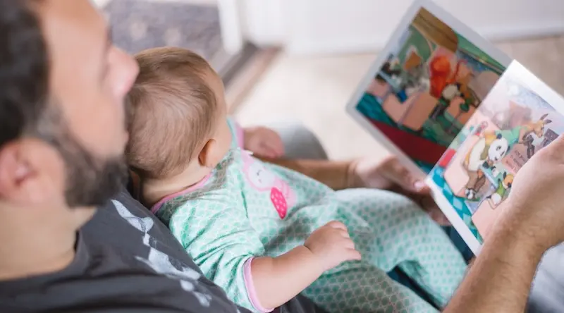 A man and a baby are sitting together, enjoying a colorful children's book. The baby is wearing a patterned onesie. The scene is warm and nurturing.