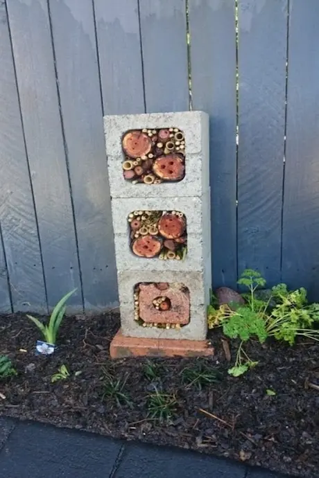 A vertical stack of cinder blocks filled with bamboo and circular wood pieces serves as a bee hotel. It's set against a wooden fence in a garden bed.