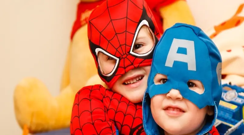 Two children are smiling, dressed as superheroes. One wears a red and black Spider-Man costume, the other a blue Captain America outfit. They look happy and playful.