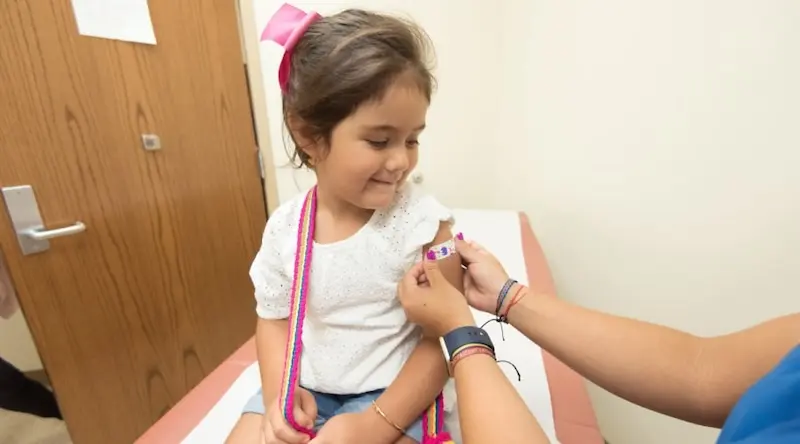 A young girl sits on a medical exam table, smiling as a healthcare worker applies a colorful bandage to her arm. The scene is warm and reassuring.