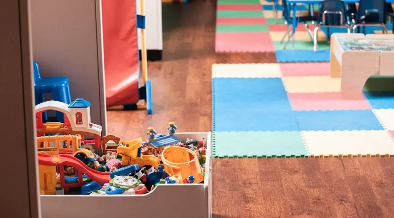 A colorful preschool classroom with a box of assorted toys like blocks and figurines on a wooden floor. Foam mats and small blue chairs fill the background.